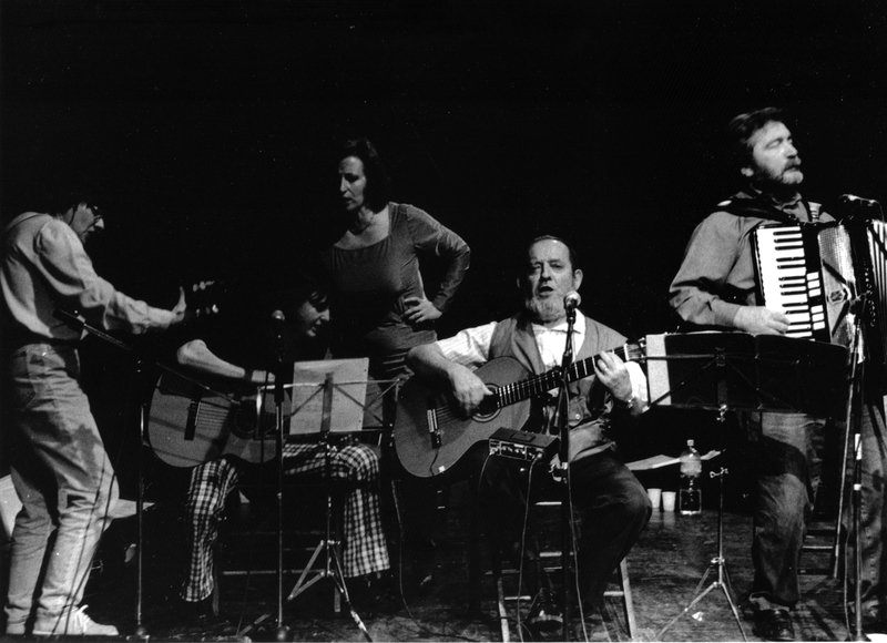 InCanto 1999 - Il nuovo canzoniere italiano - Foto di Angela Chiti che ritrae Fausto Amodei seduto su una seggiolina di legno su un palco, mentre canta accompagnandosi con la chitarra, insieme ad altre e altri sul palco insieme a lui, davanti a uno sfondo nero.