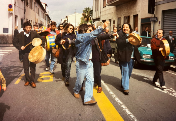 Una foto che ritrae la testa di un piccolo corteo di suonatori festanti che camminano lungo una via di Sesto Fiorentino; tra gli altri c'è Marcello Colasurdo che, sorridendo, suona un tamburello con sonagli.