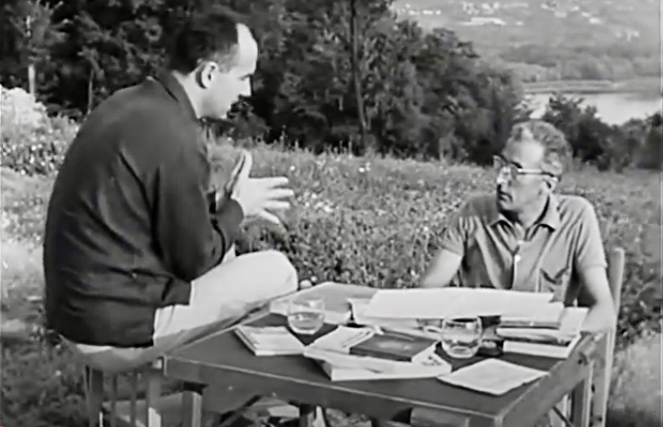 Un fotogramma dal documentario in bianco e nero «Dentro “A floresta”», che mostra Luigi Nono e Giovanni Pirelli mentre discorrono seduti su due sedie davanti a una piccola scrivania piena di libri e carte, con due bicchieri d'acqua, uno semipieno e uno vuoto, in un prato montano davanti a una boscaglia fitta, declinante verso un laghetto.